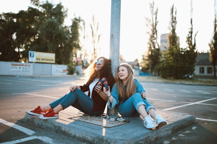 Kaksi tyttöä istuvat nojaten tolppaan Coca cola -pullot kädessään ja nauravat.