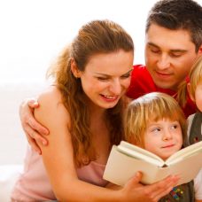 Äiti, isä ja kaksi lasta istuvat sohvalla, äiti lukee lapsille kirjaa