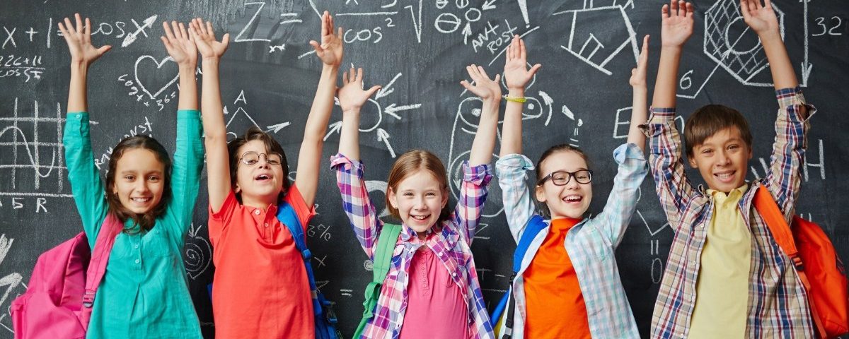 Viisi lasta seisoo koululuokassa liitutaulun edessä iloisina ja kädet pystyssä