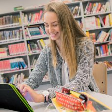 Opiskelija opiskelee hymyillen kirjastossa. / Student smiling and studying in a library.