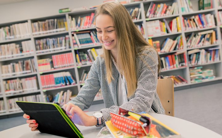 Opiskelija opiskelee hymyillen kirjastossa. / Student smiling and studying in a library.