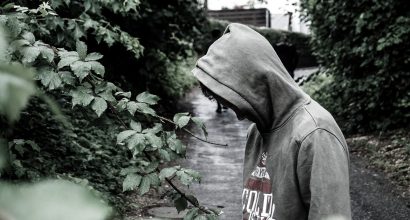 Huppupäinen nuori katsoo alaviistoon synkän sävyisessä kuvassa. An adolescent with a hood looks down in a darkly toned image.