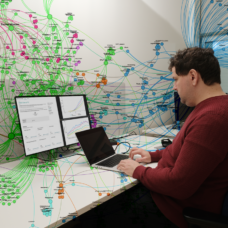 Tutkija tietokoneen ääressä, tietokoneen ruudussa datapankki, seinälle heijastunut kuva tilastoista muodostetusta kuvasta.