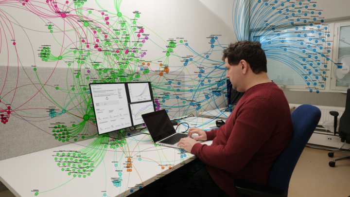 Tutkija tietokoneen ääressä, tietokoneen ruudussa datapankki, seinälle heijastunut kuva tilastoista muodostetusta kuvasta.